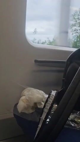 The video shows the view from inside a train. A black backpack leans against the side of the train seat. Next to the backpack is a small pile of crumpled white paper, possibly tissues. Through the train window, one can see a cloudy sky and some trees. The overall setting suggests a train journey, possibly during the day, with a focus on the immediate interior of the train car.