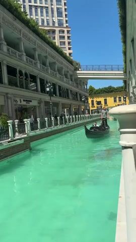 A black gondola boat with three occupants glides on a bright turquoise canal under a clear blue sky. A man stands at the stern, propelling the boat with a long pole, while two passengers are seated in front of him. The canal is flanked by multi-story buildings with balconies and shops on the lower levels, resembling a classical European style. A tall, modern high-rise building is visible in the background to the left. Further down the canal, a pedestrian bridge crosses over, connecting the buildings on either side. A yellow building is visible in the distance on the right side of the canal.