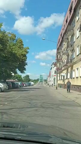 The video shows a view from inside a car driving down a street in a European city on a sunny day. On the left side of the street, there are parked cars and a tree. On the right side of the street, there are buildings, one of which appears to be an apartment building with balconies. In the distance, there is a green bridge. There are a few people walking on the sidewalk on the right side of the street. The sky is blue with white clouds.