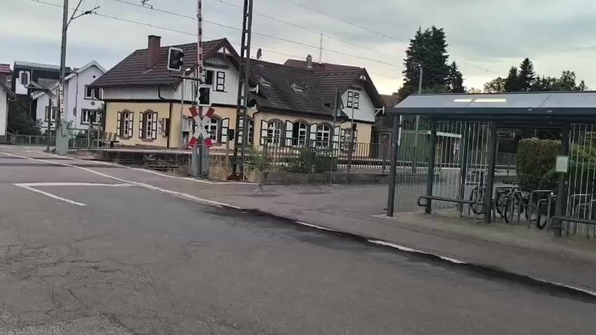 A train crossing with a red and white barrier arm is visible. A small building with yellow walls and white trim, likely a train station, sits behind the crossing. A bus stop shelter with a metal fence and parked bicycles is to the right of the station. The sky is overcast.