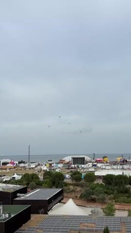 The video shows an aerial view of a coastal area with an event setup. Several small planes are flying in formation, leaving contrails in the sky, suggesting an air show or similar performance. Below the sky, a body of water, likely the sea, is visible. Along the coastline, there are numerous tents, structures, and stages, indicating a festival or large gathering. In the foreground, there are buildings with solar panels on their roofs, and some trees. The sky is overcast, with a muted, greyish hue.