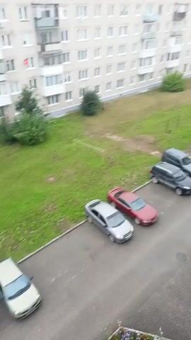 This is a still shot from a video taken from a high vantage point, looking down on a residential area and a parking lot. In the foreground, a balcony railing is visible on the right side. Below, a paved area is filled with parked cars. A light-colored sedan is in the front left, followed by a dark red sedan, and then two SUVs, one dark gray and one black. Beyond these, more cars are parked in a line along a paved road. In the background, a tall, light-colored apartment building stands on the left, with green grass and trees between it and the parking lot. Further back, there are more buildings, some appearing to be commercial, and a treeline. The sky is overcast. The cars are stationary, and no people are visible in the immediate vicinity of the parking lot.