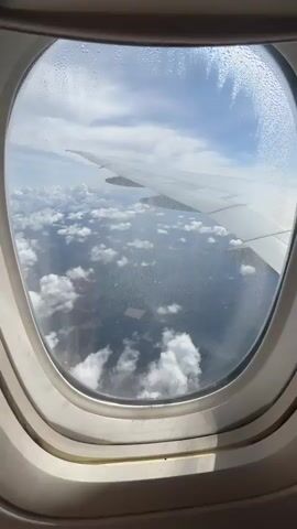 The video provides a view from an airplane window, looking down at the landscape below. The window has water droplets on the outside, creating a slightly obscured view. A portion of the airplane wing is visible in the upper right of the frame. Below, fluffy white clouds are scattered across the sky, casting shadows on the ground. The landscape appears to be a mix of dark green forested areas and lighter colored, possibly agricultural, fields. A distinct rectangular field, lighter in color than its surroundings, is visible. The overall scene suggests the plane is in flight over a rural or natural area.