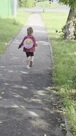 A young child walks away from the camera down a paved path. The child has blonde hair and is wearing a pink jacket, black shorts, and white shoes. The child is carrying a round pink backpack with a cartoon character on it, and a bottle of water in their left hand. The path is bordered by grass on both sides, and there are trees and bushes visible in the background. The sky is overcast. The scene appears to be in a suburban or park-like setting.