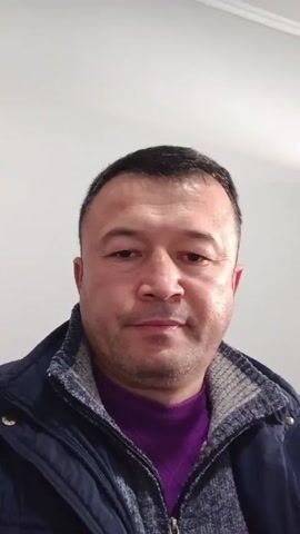 The video features a man in a close-up headshot. He has short dark hair, slightly receding at the temples, and a light stubble beard. He is wearing a dark blue jacket with a gray patterned collar and a purple sweater underneath. The background is a plain white wall. At one point, the man's finger comes into view in the foreground.