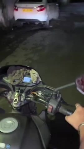 A first-person perspective video captures a motorcycle rider's point of view as they navigate a dark, paved road at night. The motorcycle's handlebars and instrument panel are visible in the foreground, with a digital display showing numbers. A white car is parked on the left side of the road. Further down the road, trees line the sides, and streetlights illuminate the path. Several people are walking in the distance, appearing as blurry figures due to the low light conditions. The environment suggests a residential or semi-urban area at nighttime.