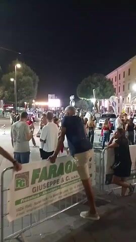 A crowd of people are gathered on a street at night. There are temporary barriers set up, possibly for an event. In the foreground, a man in a blue shirt and beige shorts is leaning over a barrier. In the background, buildings and streetlights are visible. The atmosphere appears to be festive.