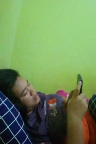 The video is a close-up, blurry shot of a person lying down on a bed, looking at their phone. The person is wearing a purple shirt and has dark hair. They are holding a black phone in their right hand. The background is a lime green wall. There is a blue and white checkered pillow under the person's head. The lighting is dim.