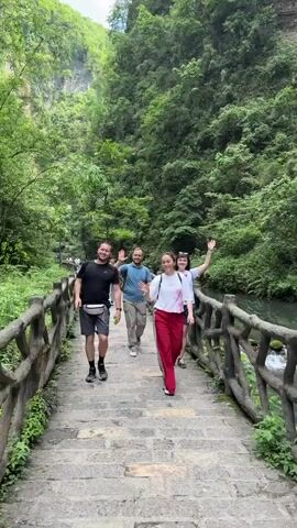 Four adults, three men and one woman, walk along a paved path through a lush, green canyon. A low, rustic wooden fence borders the path on one side, with a small stream flowing just beyond. Two of the adults wave and look directly at the camera, and the man at the front of the group makes a fist. The men are wearing casual clothes and the women are wearing light colored t-shirts with pants or shorts. The path leads toward a dense, green forest.  Sunlight filters through the trees, creating a bright, natural light. The overall atmosphere is cheerful and adventurous.