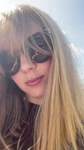 A close-up video shows a young woman with long blonde hair and bangs, wearing dark sunglasses and a dark top. Her hair is being blown around by the wind, obscuring parts of her face. She is outdoors, with a bright sky visible in the background. The video appears to be a selfie, with the camera held at a close distance to the subject.