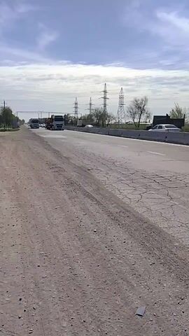 The video shows a highway with light traffic. A flatbed truck hauling an excavator passes by. The road surface is cracked and there is dirt and gravel on the shoulder. Power lines are visible in the distance. The sky is partly cloudy.