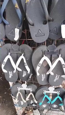 The video opens with a close-up view of a rack of black flip-flops. The camera then pans to show more racks of flip-flops in various colors, including blue and gray. The scene then shifts to an outdoor market stall selling shoes. Several people are visible in the background, and a variety of shoes are displayed on shelves and tables. The camera then moves to a wider shot of the market, showing rows of shoes packaged in plastic bags. Motorcycles are parked nearby, and a few people are browsing the merchandise. The video concludes with a view of the street in front of the market, with traffic and pedestrians visible.
