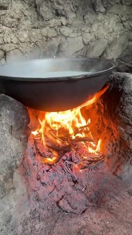 A large metal pot filled with a white liquid sits over an open fire built in a stone structure. Flames from the fire lick the bottom of the pot, causing the liquid to steam. The fire is made of wood and embers. The background is a rough, stone wall. The scene is dimly lit, with the fire providing the primary light source.