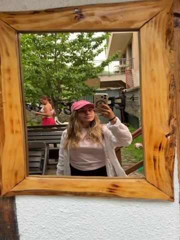 A young woman with blonde hair, wearing a pink baseball cap, a white open shirt over a pink t-shirt, and black pants, takes a selfie in a rustic wooden mirror. The mirror is framed by natural, unfinished wood with visible grain and knots. In the reflection, behind her, another woman is visible, wearing a pink sports bra and pink shorts, standing near some outdoor seating and greenery. The scene appears to be an outdoor setting, possibly a backyard or patio, with trees, plants, and a building in the background. The overall atmosphere seems casual and relaxed. The time of day is difficult to determine precisely, but the lighting suggests it could be daytime, possibly overcast given the soft light. The setting with the rustic mirror and outdoor elements could be in a more rural or natural area. No visible text is present in the image.
