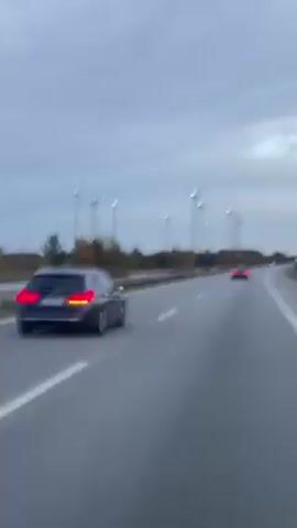 A car travels on a highway. There are other cars ahead on the road and many light posts and some trees to the side. The sky is gray. The road is gray, and the painted lines are white. The video is shot from behind, with a blurry background.