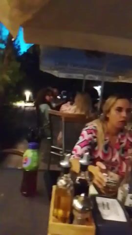 The video begins with a close-up of a woman with blonde hair sitting at an outdoor table at night. She is wearing a colorful, geometric-patterned shirt and looking to her right. In front of her on the table are several bottles and a glass of beer. The camera then pans to the left, showing a plate of food, an ashtray, and another glass with a dark liquid and a slice of lemon. Further to the left, a bottle of Coca-Cola and a dessert in a glass are visible. The background shows other tables with people seated, dimly lit by overhead lights and outdoor lamps. Umbrellas are set up over some of the seating areas. The overall atmosphere appears to be that of a casual outdoor dining or bar setting at night. The camera continues to pan right, showing more tables and people in the background. The video ends showing a broader view of the outdoor seating area with more patrons visible in the distance.
