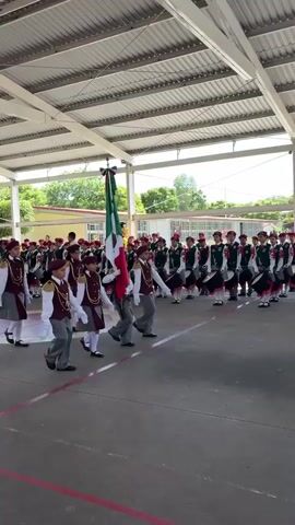 A group of young people in matching uniforms, consisting of grey pants, maroon vests with gold trim and epaulets, white shirts, and maroon hats with gold trim, are marching in formation. Some are carrying drums and wearing white gloves. The central figures in the foreground are holding a large Mexican flag. They are marching in an outdoor area with a covered roof overhead. In the background, more individuals in similar uniforms are visible, some playing drums. There are buildings with yellow and white walls and red roofs visible in the distance. The ground is a reddish-brown paved surface with white lines marking boundaries. The overall scene appears to be a school parade or ceremony.