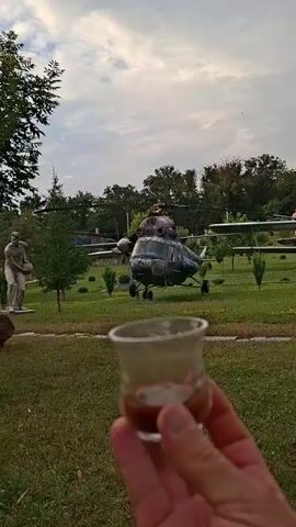 A close-up shot shows a person's hand holding a small glass containing a dark liquid, possibly coffee or a drink. The background is an outdoor setting with a grassy area, trees, and a statue on the left. In the middle ground, a helicopter is prominently displayed, and other helicopters are visible in the distance. The sky above is overcast. The camera appears to be positioned at eye level with the hand holding the glass.