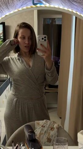 A young woman with short brown hair, wearing a grey loungewear set and a gold pendant necklace, records herself in a mirror with her phone. She stands in what appears to be a bedroom or living area, striking various poses, adjusting her hair, and making a slight pouty face. The mirror is framed with a decorative feather pattern and has an LED light strip along its top edge. Behind her, reflections show a room with a baby cot, a window or door with light-colored curtains, and a piece of furniture. A chair with a patterned cloth (featuring rabbits) sits in the foreground. The lighting in the room is warm, suggesting an evening or indoor setting.