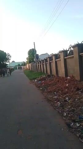 This video shows a street scene with a paved road on the right side of the frame and a large pile of debris and trash along the side of the road. A tall, ornate fence runs along the right side of the road, topped with what appears to be barbed wire. Further down the road, a group of people is walking away from the camera, and a building with a green roof is visible in the distance. On the left side of the road, there is a large tree and some smaller structures. The sky is clear and appears to be late afternoon or early morning. The overall impression is one of a somewhat neglected urban or semi-urban environment.