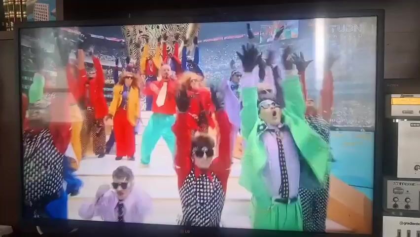 The video shows a wide shot of a television screen displaying a performance. A group of people in colorful, retro-inspired outfits are dancing and singing on a stage. The lead singer, a bald man in a red shirt and teal pants, is holding a microphone. The background appears to be a stadium filled with an audience, though they are out of focus. The performance features bright lighting and energetic movements. The overall scene suggests a lively and engaging musical event.