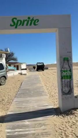 The video shows a sandy area, possibly a beach or desert landscape, with a wooden boardwalk leading into it. At the start of the video, a dark-colored vehicle is seen driving on the sand in the distance. The camera is positioned near an entrance or gateway marked with Sprite branding. The sky is clear and blue. The camera pans slightly to the right, showing more of the sandy expanse. The vehicle continues to move in the distance, leaving tire tracks on the sand. In the background, there are dunes or sand hills. A contrail from a plane can be seen in the sky. A few people appear small in the distance.