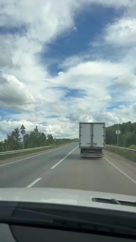 The video captures a view from inside a vehicle traveling on a highway. The perspective is looking forward through the windshield. The road ahead is paved with asphalt and marked with white lines. On the right side of the road, a large white truck with a trailer is visible, also traveling in the same direction. Beyond the road and to the left, there is a grassy shoulder with some trees and vegetation. A white guardrail runs along the edge of the road. In the distance, more trees and fields can be seen. The sky is blue with scattered white clouds, some of which are large and fluffy. The overall impression is of a daytime journey on a rural or semi-rural highway.