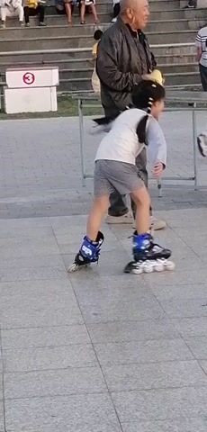 An adult male stands with a yellow cloth in his left hand, facing left. He is wearing a dark jacket, grey pants, and beige Crocs. To his right, a young girl is rollerblading. She is wearing a white shirt with grey and blue stripes, grey and white striped shorts, and blue rollerblades. She is looking to her left. Behind her, another young girl is also rollerblading, wearing a green tracksuit with white trim and white rollerblades. She is looking to her right. In the background, people are sitting on stone steps. The ground is paved with large, light grey tiles. Two metal tables are positioned in front of the girls. The overall lighting suggests it is daytime, likely late afternoon due to the soft sunlight.