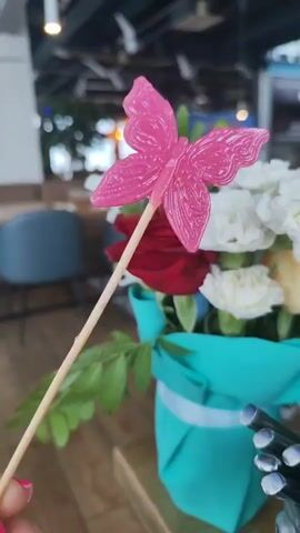 A close-up, shallow depth-of-field shot shows a pink butterfly-shaped lollipop on a wooden stick being held in front of a floral arrangement. The lollipop has white swirl detailing on its wings. The floral arrangement is in a teal colored wrapping and contains red roses and white carnations. The background is blurred, showing hints of a restaurant or cafe setting with wooden tables and chairs.