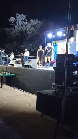 The video shows an outdoor stage at night. There are several people on the stage. Two people are standing in the center of the stage, appearing to be singing or performing. One is a woman in a long, dark dress, and the other is a person in a sparkly outfit. To the left of them, a person in a white shirt is standing near a laptop and some equipment. To the far left, another person is seated at what appears to be a keyboard. On the right side of the stage, another person in a suit is standing. There are speakers set up in front of the stage. The background shows trees and lights. There is a banner visible on the stage with some text and graphics. The overall scene appears to be a live performance or event.