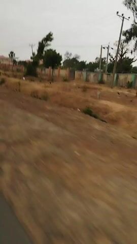 The video shows a moving vehicle (possibly a motorcycle) driving on a paved road. There are several other motorcycles on the road ahead, traveling in the same direction. A large truck can be seen further in the distance. The environment includes dry land, trees, and buildings on either side of the road. Streetlights are also visible along the road. The sky is overcast.