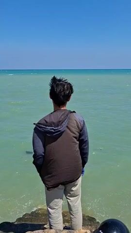A person stands with their back to the camera, looking out at a vast, calm expanse of greenish-blue water under a clear blue sky. The person is wearing a dark vest over a blue long-sleeved shirt and light-colored pants. The water stretches to the horizon, where it meets the sky in a faint line. The foreground shows a rough, textured surface, possibly a dock or walkway, with some pebbles and debris visible. A black object, possibly a helmet, is partially visible in the bottom right corner of the frame. The overall scene evokes a sense of tranquility and openness.