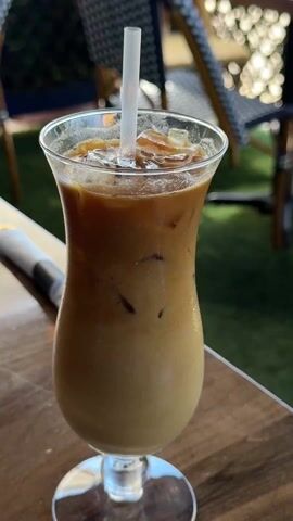 A close-up shot displays an iced coffee in a tall, tulip-shaped glass, garnished with ice and a white straw. A hand with manicured nails stirs the drink with the straw. The drink appears to be a mix of coffee and milk or a cream, with a lighter hue at the bottom and a darker layer at the top. The glass rests on a wooden table. A silver-wrapped utensil is partially visible on the left side of the frame. In the background, there is blurred greenery and outdoor patio furniture, suggesting the scene is set in an outdoor cafe or restaurant setting.