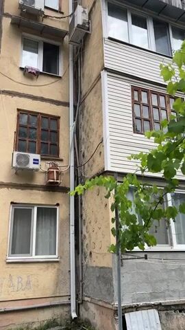 This media file shows a close-up view of the exterior of an apartment building in Pitsunda, Abkhazia. The building appears to be older, with a textured, yellow-toned facade and some areas of concrete. There are multiple windows visible, some with wooden frames and others with modern white frames.

Prominently featured is an air conditioning unit on the left side of the frame, with the "Haier" brand visible. Below it, a rusty, cylindrical object, possibly a gas cylinder, is mounted in a wire cage. A white downspout runs vertically along the corner of the building, and several electrical wires are routed along the exterior.

On the right side of the building, a section is covered with white siding, and another wooden-framed window is visible. A vine with green leaves is growing up the side of the building, partially obscuring some of the details. The lighting suggests it is daytime, and the weather appears to be overcast or at least not brightly sunny, as there are no strong shadows.

The overall impression is one of a lived-in, slightly weathered urban environment. The juxtaposition of older architectural elements with modern additions like the air conditioner and siding, along with the natural element of the vine, creates a visually interesting scene. The image appears to be a still shot, with no discernible activity or event taking place beyond the static view of the building's facade. No people are visible in the media file. There is some faint, illegible text on the lower left portion of the building's wall.
