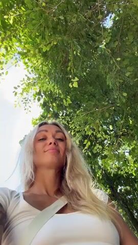 A fair-skinned woman with long blonde hair and a shoulder tattoo takes a selfie in front of a white and gold pagoda-like structure under a bright blue sky. She is wearing a white top and a white bag strap is visible. Lush green trees are present, and a small pond is visible in the lower right corner of the frame. The camera angle is low, pointing up at the woman.
