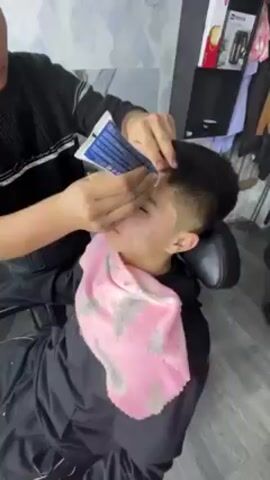 A man sits in a black barber's chair with a pink towel draped around his neck. His hair is short, black, and neatly styled. A person wearing a black sweater applies a white cream from a blue and white tube onto the man's face. The room has light blue and white textured wallpaper. There's a black storage unit visible in the background, displaying various products.