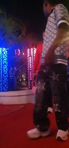 The media file captures a nighttime event at Bar Jirgi Summa, Nigeria, likely a social gathering or celebration. The scene is illuminated by ambient lighting, with some areas featuring blue and yellow lights. A red carpet is laid out, suggesting a VIP or special occasion.

The primary subjects are people, with a focus on two individuals in the foreground. A person in a black sequined dress, holding a blue, ruffled garment, is seen from the waist down, moving with a slight sway. Beside them, a person in a black patterned shirt and sparkling black pants walks past. Further in the background, another individual is visible, wearing a red jacket with "AMIRI" emblazoned on the back, accompanied by a tan handbag.

The activity appears to be people arriving or moving through the venue. The atmosphere seems festive, with the subjects appearing engaged and perhaps in good spirits. The time of day is clearly night.

Notable details include the shimmering fabric of the clothing, the ornate decorative screens in the background, and the presence of what looks like a security guard or staff member seated on the right. The "AMIRI" text on the red jacket is a prominent detail. The overall impression is of an upscale, lively evening event.