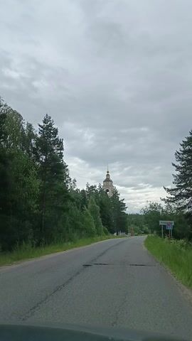 This media file shows a road with trees on either side and a cloudy sky. In the distance, a church steeple with a golden dome is visible above the trees. A sign with Russian text that appears to read "ГРИБЧИХА" (Gribchiha) is also visible on the right side of the road. The scene suggests a rural or semi-rural setting in Russia. The weather appears to be overcast, and the time of day is likely daytime. The video captures a journey along the road, with the church steeple remaining a consistent visual element in the background. There are no people visible in the footage. The overall mood is peaceful and scenic, characteristic of a drive through the countryside.
