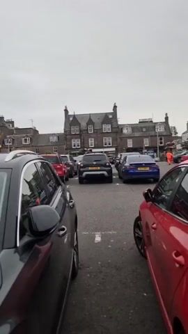 The video pans across a parking lot with several cars parked. In the background, there are buildings with distinctive architecture, including a tower with a clock. There is also a large white bus with "COACHES" written on its side. A person wearing a maroon jacket and black pants is standing in the parking lot, looking towards the cars. The sky is overcast. The footage appears to be taken on a slightly damp day, as indicated by reflections on the car surfaces.