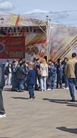 The video shows a public square in Russia on a sunny day, likely celebrating Victory Day on May 9th, as indicated by the stage banner with "С Днем Победы! 9 МАЯ" (Happy Victory Day! May 9th). People are walking around, some grouped in what looks like junior military cadet uniforms. There's a stage set up with lighting and banners related to WWII and Soviet symbolism, including the hammer and sickle and a red star. Additionally, there are stalls or booths set up, and other Soviet-era flags are around.