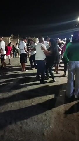 The video shows a crowd of people dancing at night.  A man and a woman are slow dancing in the center of the frame, while other couples are spread throughout the area.  Most of the people are wearing casual clothing, and there is a person sitting on the ground in the lower right corner of the frame.  The scene appears to be outdoors, with street lights providing illumination. The ground appears to be uneven and possibly unpaved.  Long shadows from the streetlights are cast across the scene. There is music playing in the background.