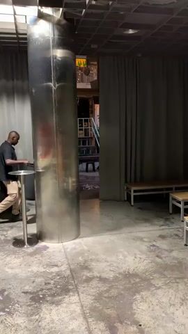 A person wearing a black t-shirt and light-colored pants walks past a large silver pillar. The person is carrying a black trash bag in their right hand and a black object in their left. The person walks behind the pillar and out of frame. The environment appears to be an indoor space with gray curtains and a concrete floor. There are two benches visible on the right side of the frame.