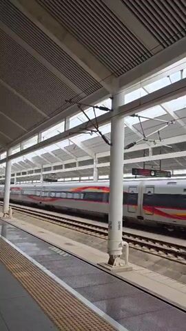 A high-speed train with a red and yellow stripe along its side speeds past a covered train platform. The platform has a textured yellow and black safety strip along the edge. Several white support columns hold up the roof structure, which is made of metal and glass panels. Overhead wires and electrical equipment are visible above the train tracks. The sky is visible through the translucent roof panels.