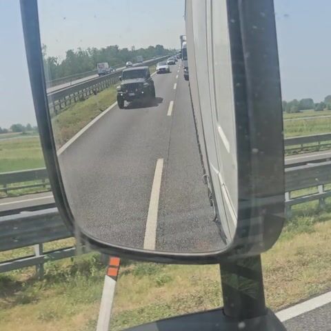 A black Jeep Wrangler is driving on a highway. The video is shot from the perspective of another vehicle, likely a truck, looking in the side mirror. The Jeep is in the right lane, ahead of the camera vehicle. The highway is asphalt with white lane markings. There are other vehicles on the highway in the distance. The environment outside the highway appears to be rural with fields and some trees. The sky is clear and blue.