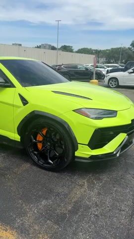 The video shows a lime green Lamborghini Urus parked in a parking lot. The car has black wheels with orange brake calipers. Other vehicles are parked in the background. The weather appears to be sunny with some clouds. The camera slowly pans along the car.