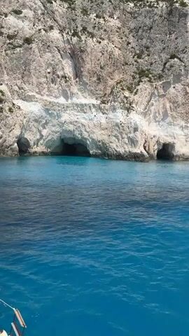 A boat is sailing in the sea. The water is turquoise and clear. To the right of the boat, there is a large cliff with several caves. The cliff is made of white rock and has some vegetation growing on it. The sky is blue and clear. The sun is shining, casting a bright light on the scene.