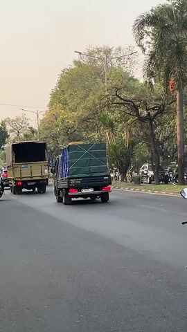 The video shows a busy street scene with several vehicles moving. In the foreground, a person on a black scooter is riding in the right lane of a paved road. The rider is wearing a black helmet and a dark jacket. Behind the scooter, two trucks are visible. The truck in the left lane is a light brown color and appears to be carrying cargo. The truck to its right is green and also appears to be carrying goods covered with a tarp. To the right of the scooter, a black pickup truck is traveling in the same direction. Further in the background, more vehicles and trees line the street. The sky is overcast or hazy. The scene appears to be in an urban or suburban area during the daytime.