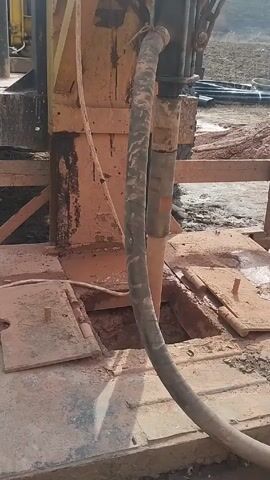 The camera captures a close-up, low-angle view of a drilling rig in operation. The rig is primarily yellow and rusty in color, suggesting it is possibly used or older.  A thick, dark grey hose is prominently featured, snaking down into an opening in the rig's platform, which is surrounded by mud and debris. The camera then tilts upwards, revealing a blue motor mounted on the upper portion of the rig. The background consists of a muddy area, some indistinct objects including pipes and a pile of dirt, and a sky with the sun peeking through the clouds. The overall impression is that the rig is set up for drilling, likely for a well or other similar excavation purpose.