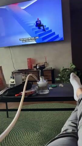 The video shows a person's legs and feet as they relax on a rug in front of a coffee table and a television. On the coffee table is a hookah with smoke coming from it. The television displays a music video with lyrics. The room is dimly lit, with the television screen and a hookah being the primary light sources.