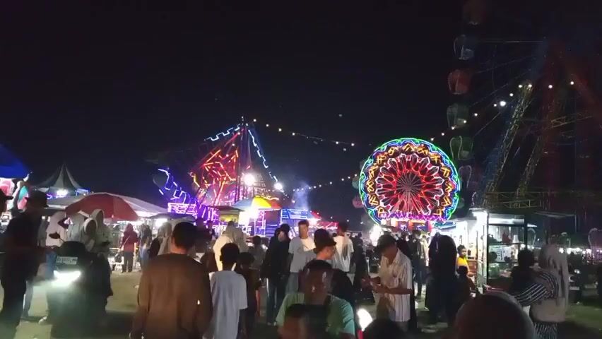 The video shows a lively night market or fair. A Ferris wheel is visible in the background, along with other illuminated rides and attractions, including one that appears to be a spinning, colorful wheel of lights and another pyramid-shaped structure with colored lights. Many people are walking around, some on motorbikes. Tents and stalls are set up, suggesting vendors are present. The overall atmosphere is festive and crowded, illuminated by artificial lights against a dark night sky.